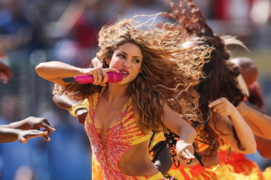 Shakira chantant au micro sur scène, éclairée par des lumières orangées lors d'un concert